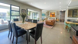 A modern living room with a dining area, a sofa, and a coffee table. There is a large window providing natural light and a view of the outdoors.