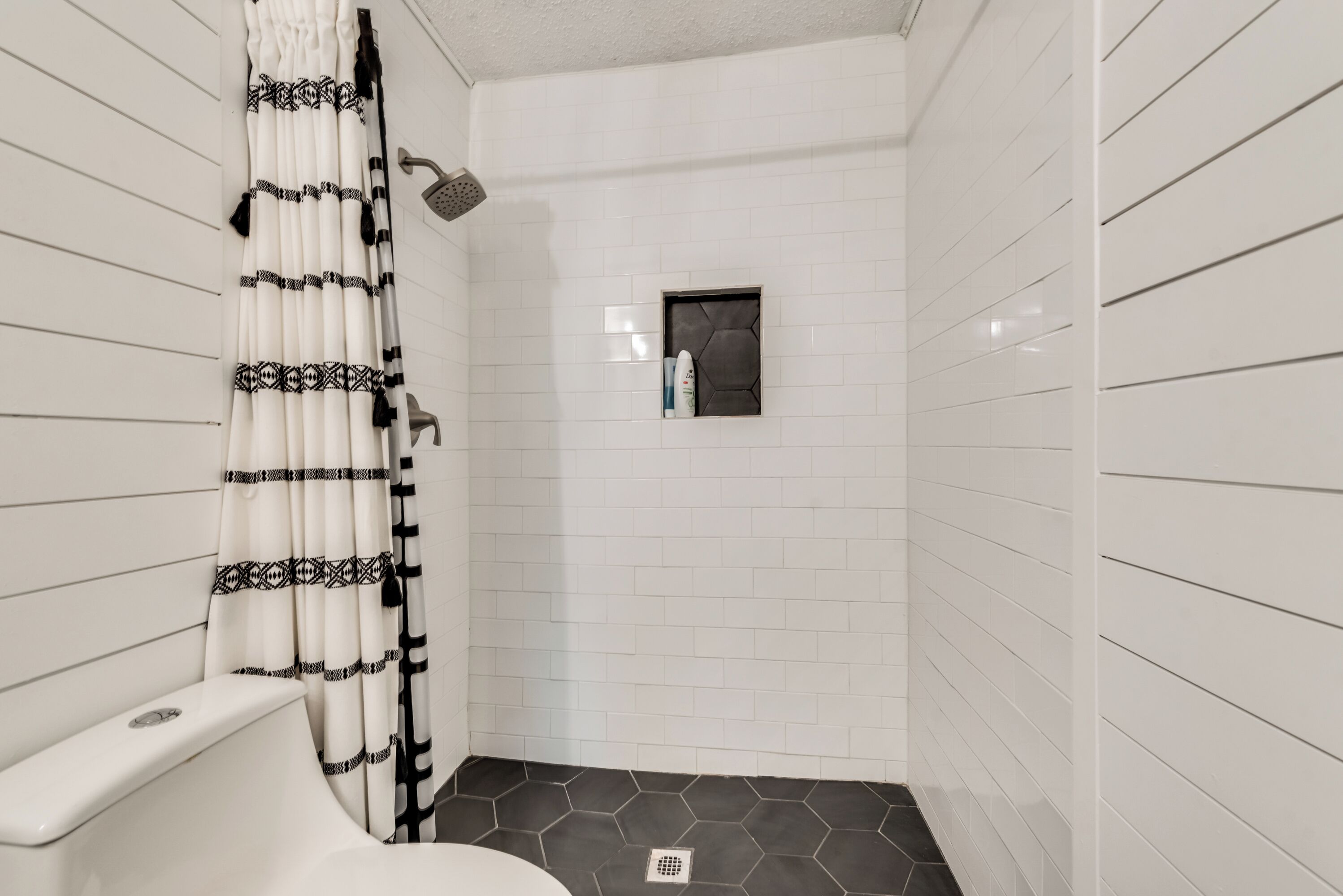 Ein Badezimmer mit Duschbereich in weißen Fliesen, Toilette und kleinem Fenster mit Duschkopf.