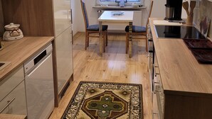 A modern kitchen with wooden cabinets, a dining table with chairs, a view of the outside through a window, and a patterned rug on the floor.