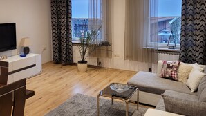 A modern living room with a flat-screen TV, a coffee table, and a sofa. There are large windows with curtains, a potted plant, and a view of the city.