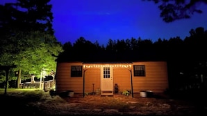 Eine kleine Holzhütte mit Veranda, beleuchtet von Lichterketten, umgeben von Bäumen.