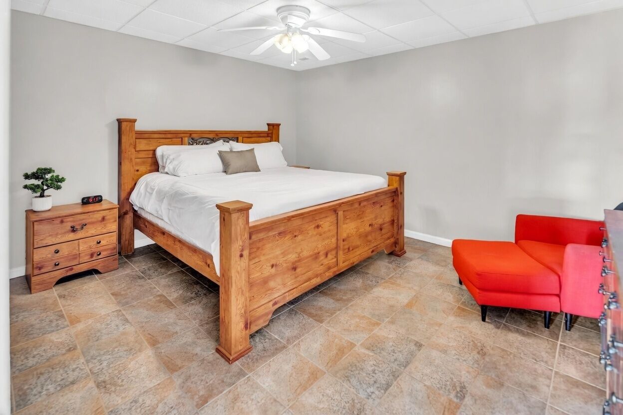 Een slaapkamer met een houten bed, een nachtkastje met lades, een rode stoel en een plafondventilator.