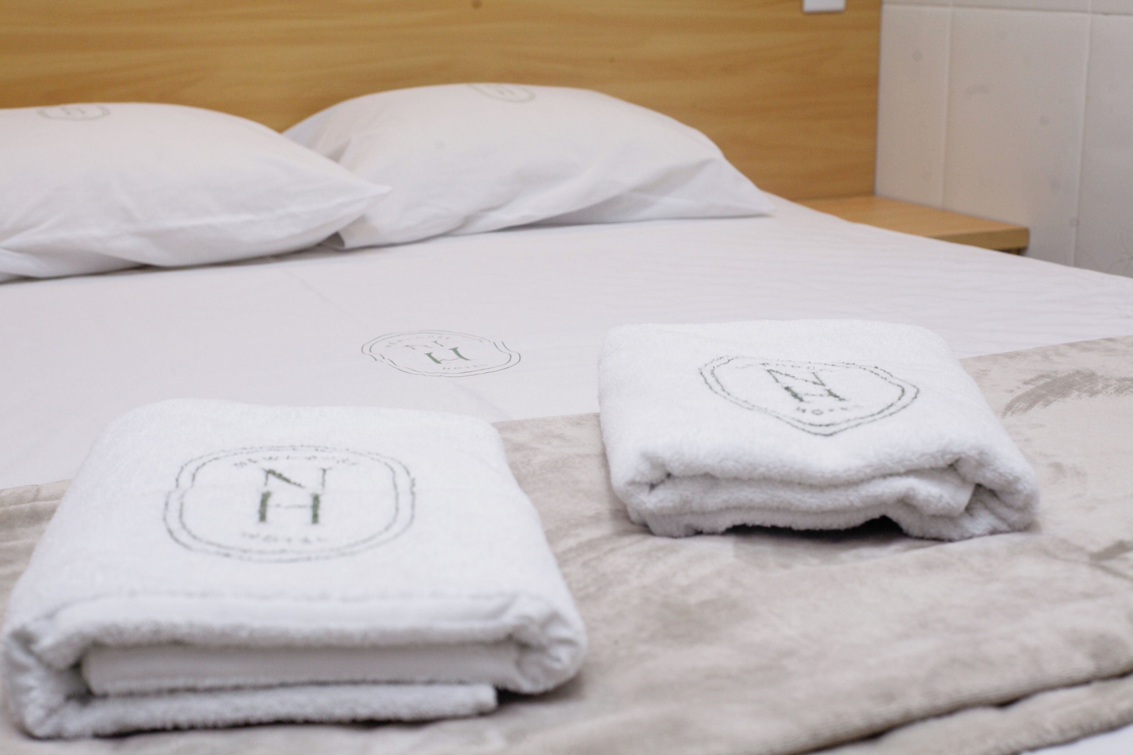 A neatly made bed with white linens and two folded towels with a monogrammed logo.