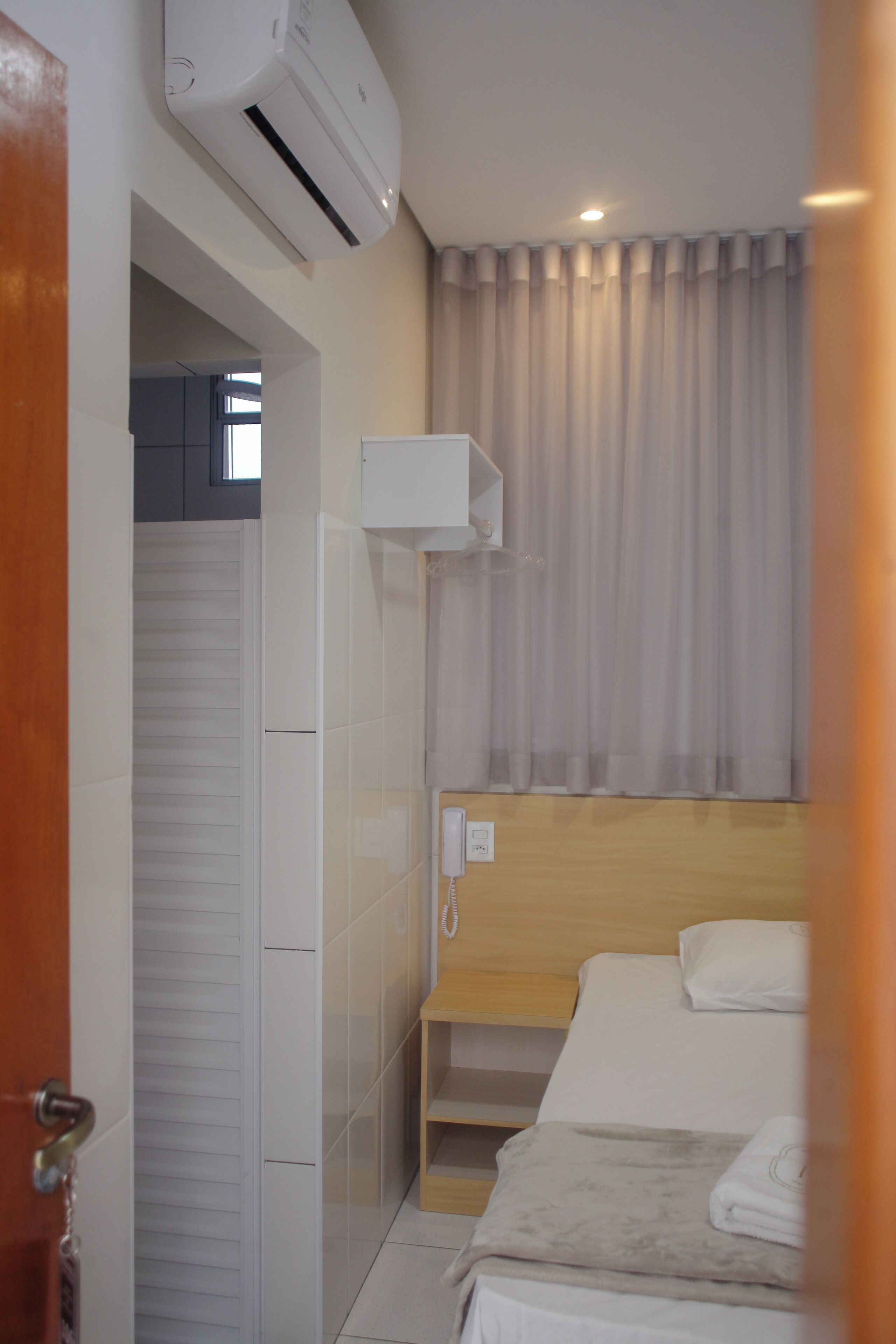 A hotel room with a bed, a wooden headboard, a bedside table, a white air conditioning unit, a window with curtains, and a door with a handle.
