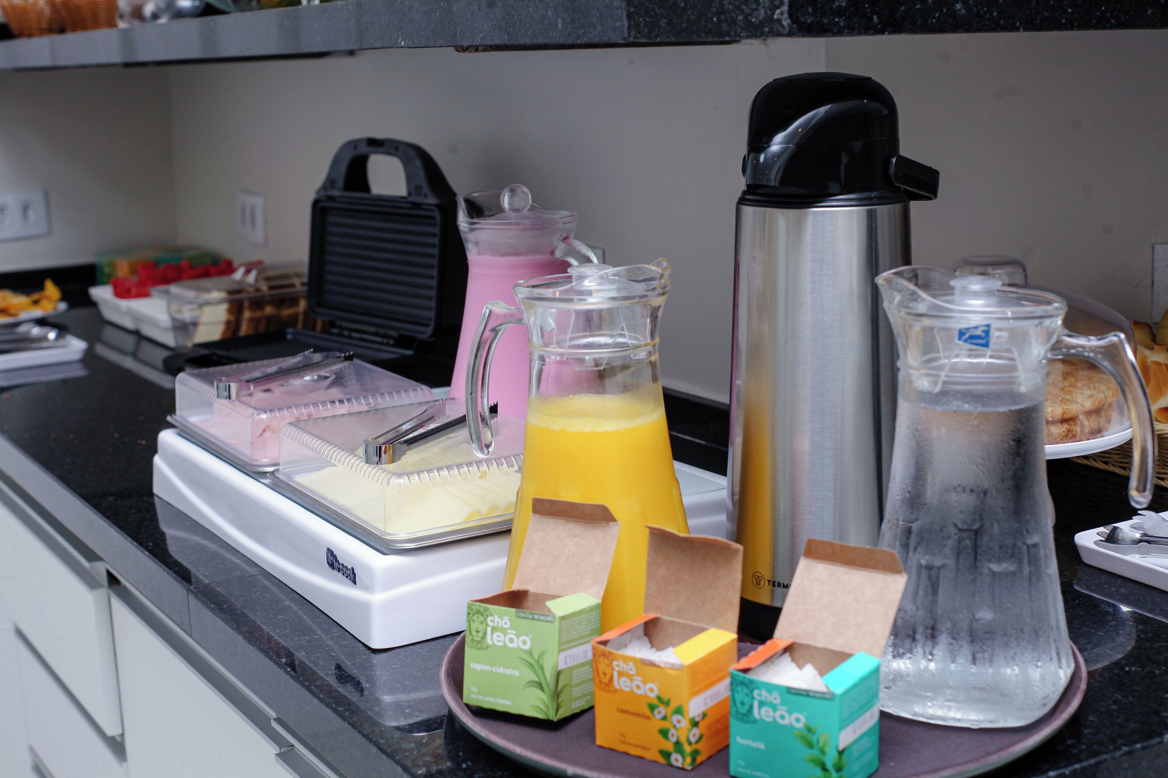 Een aanrecht met een koffiezetapparaat, een waterdispenser en een pitcher met sap. Ook staan er verschillende bakjes met eten en een bord met gerechten op het aanrecht.