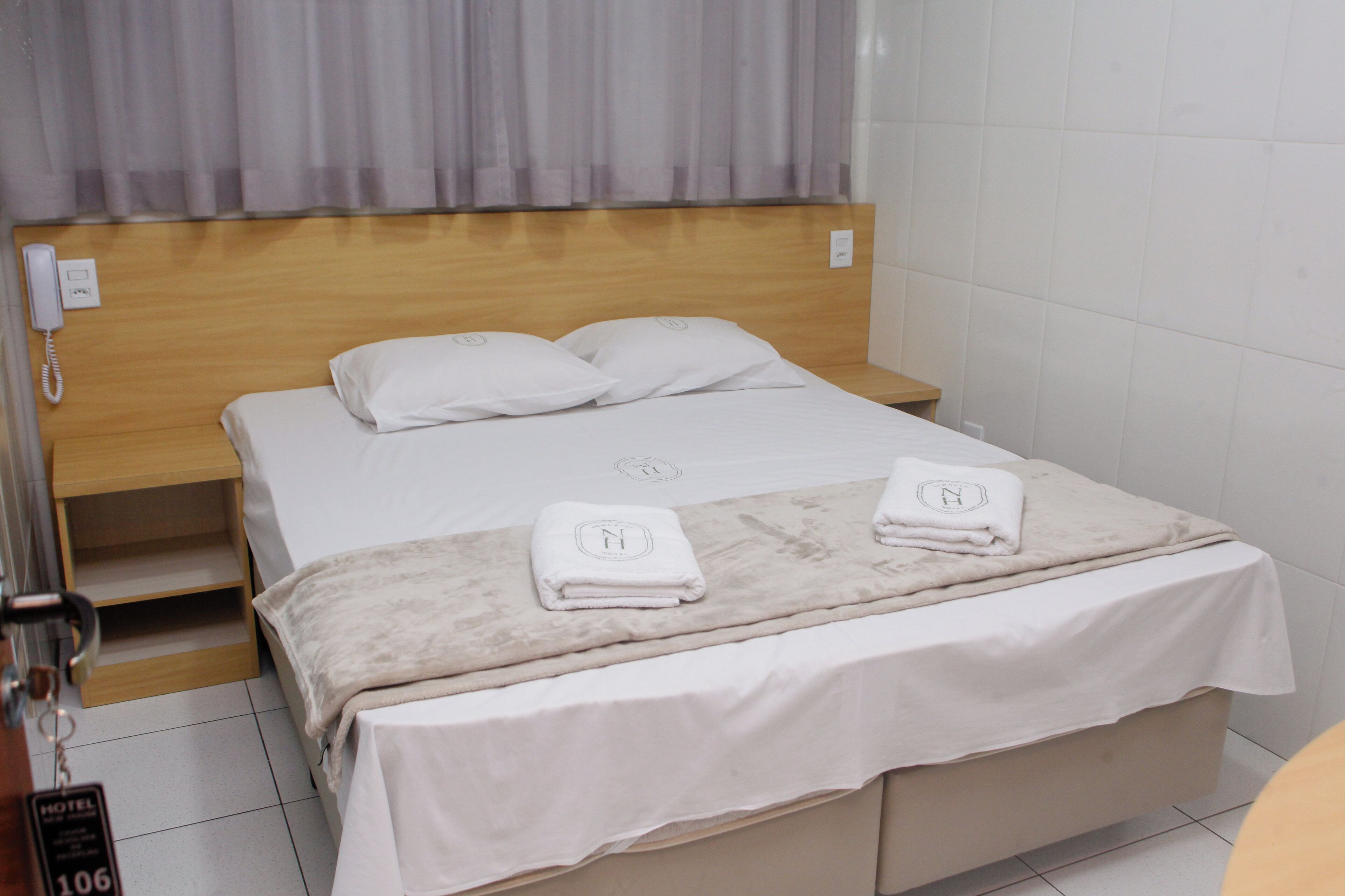 Habitación de hotel con cabecera de madera, cama con ropa de cama blanca, dos almohadas, una mesita de noche y una toalla doblada con un monograma.