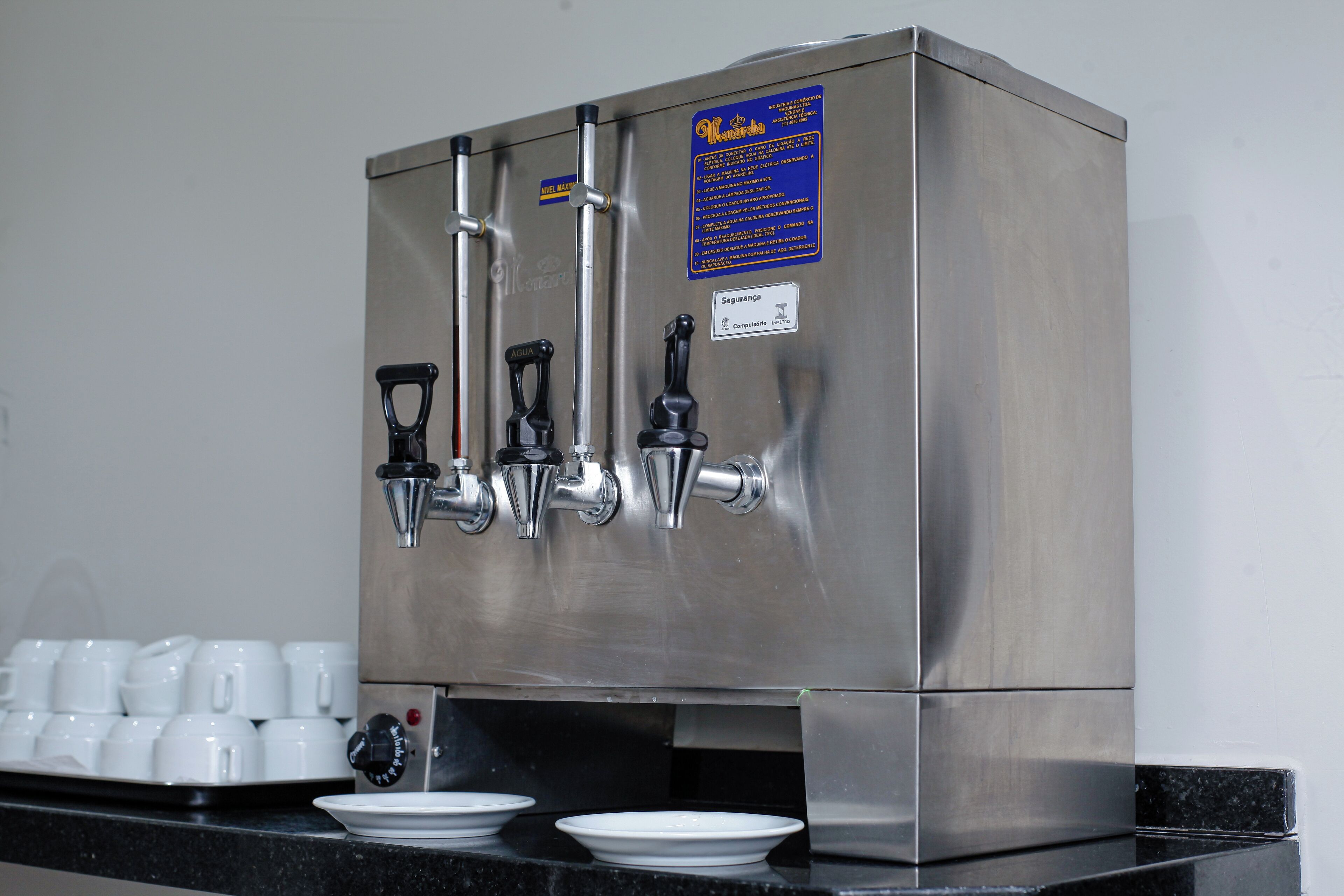 Un distributeur de boissons en acier inoxydable, doté de plusieurs robinets et d’une rangée de gobelets blancs sur le comptoir.