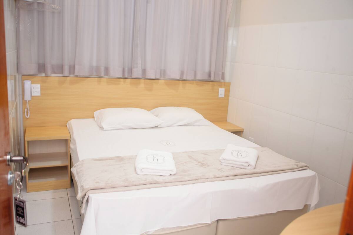 Camera d'albergo con un letto, due cuscini, una testiera in legno, un comodino e un bagno visibile attraverso una porta aperta.