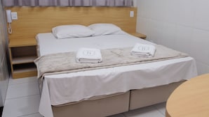 Quarto de hotel com cabeceira de madeira, cama com roupa de cama branca, duas toalhas sobre a cama, mesa de cabeceira e telefone na parede.