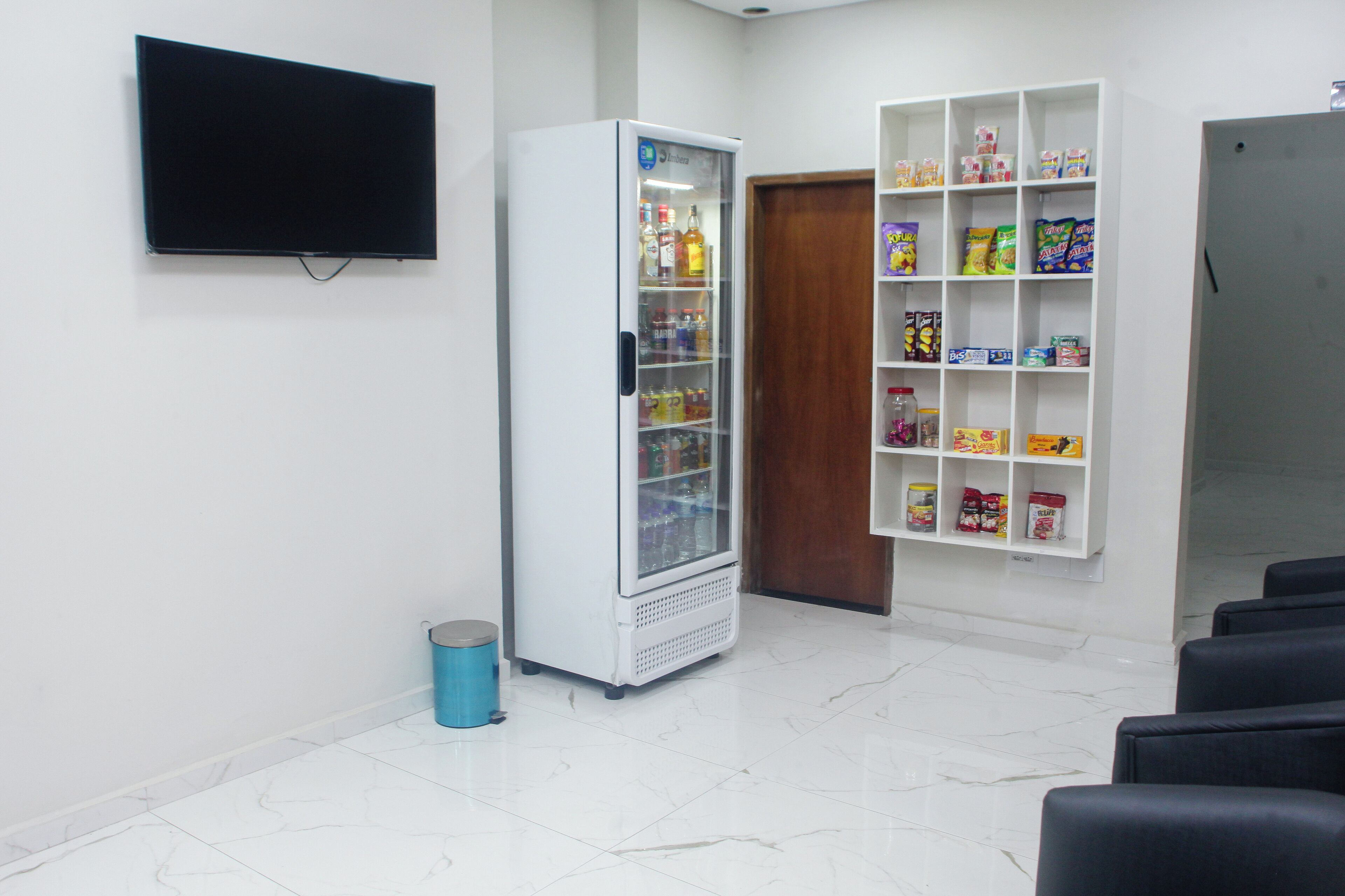 A room with a flat-screen TV mounted on the wall, a beverage refrigerator stocked with drinks, and a snack shelf with various packaged foods.