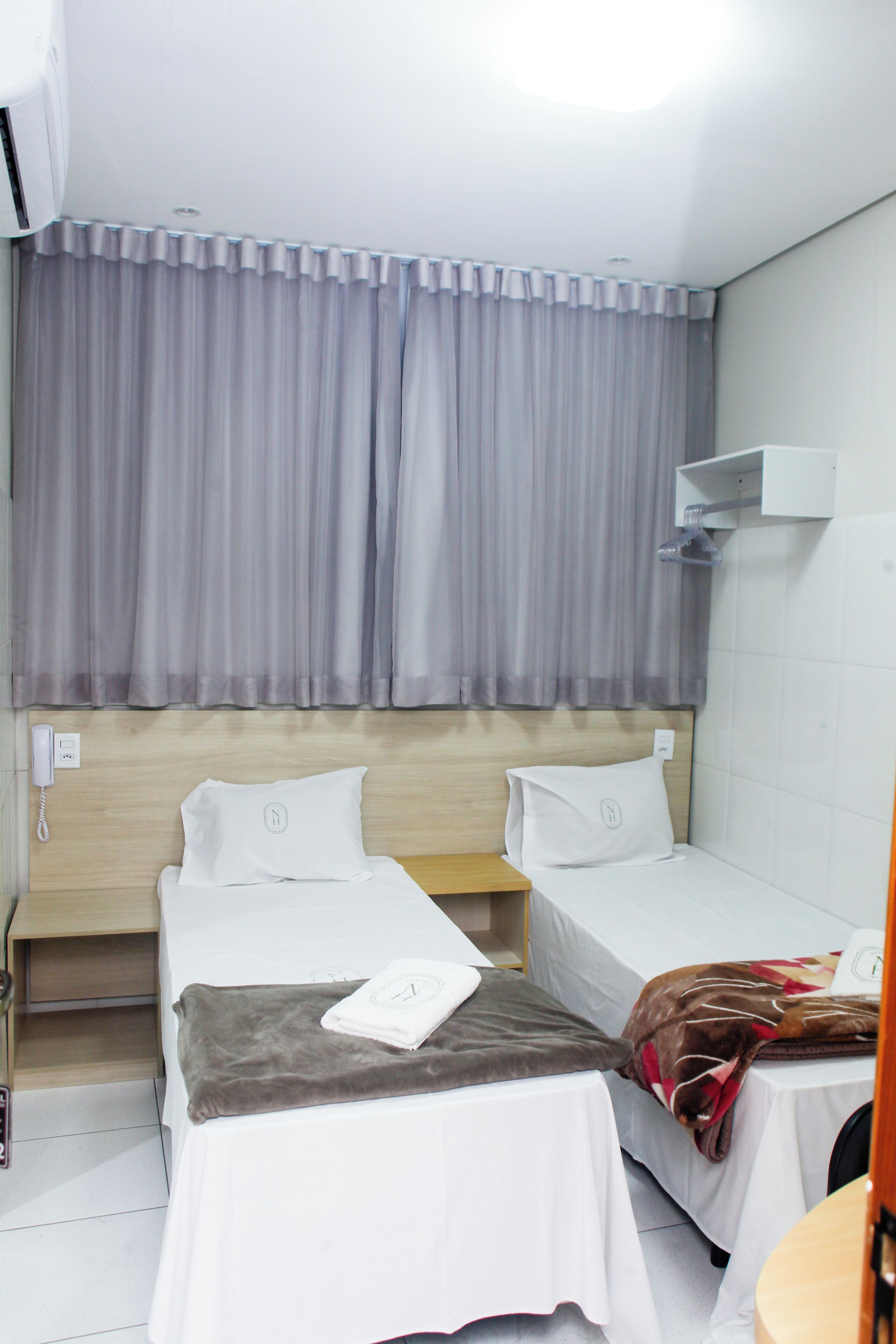 A hotel room with two single beds, a wooden headboard, a small table, and a wall-mounted shelf.