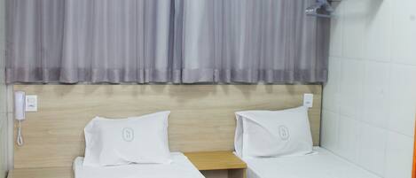 A hotel room with two single beds, a wooden headboard, a small table, and a wall-mounted shelf.