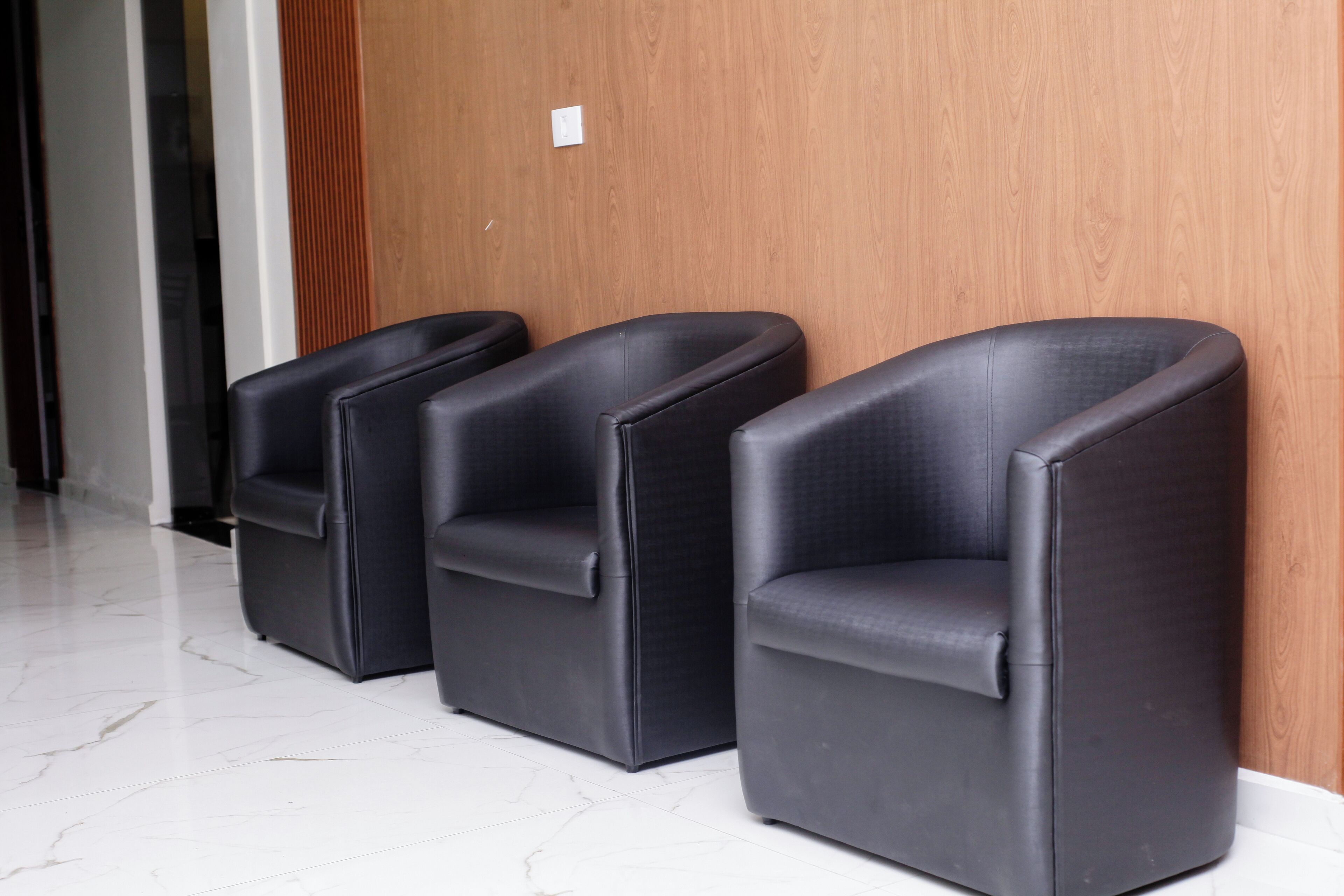 Three black leather chairs aligned against a wooden wall in a hallway with a white marble floor.