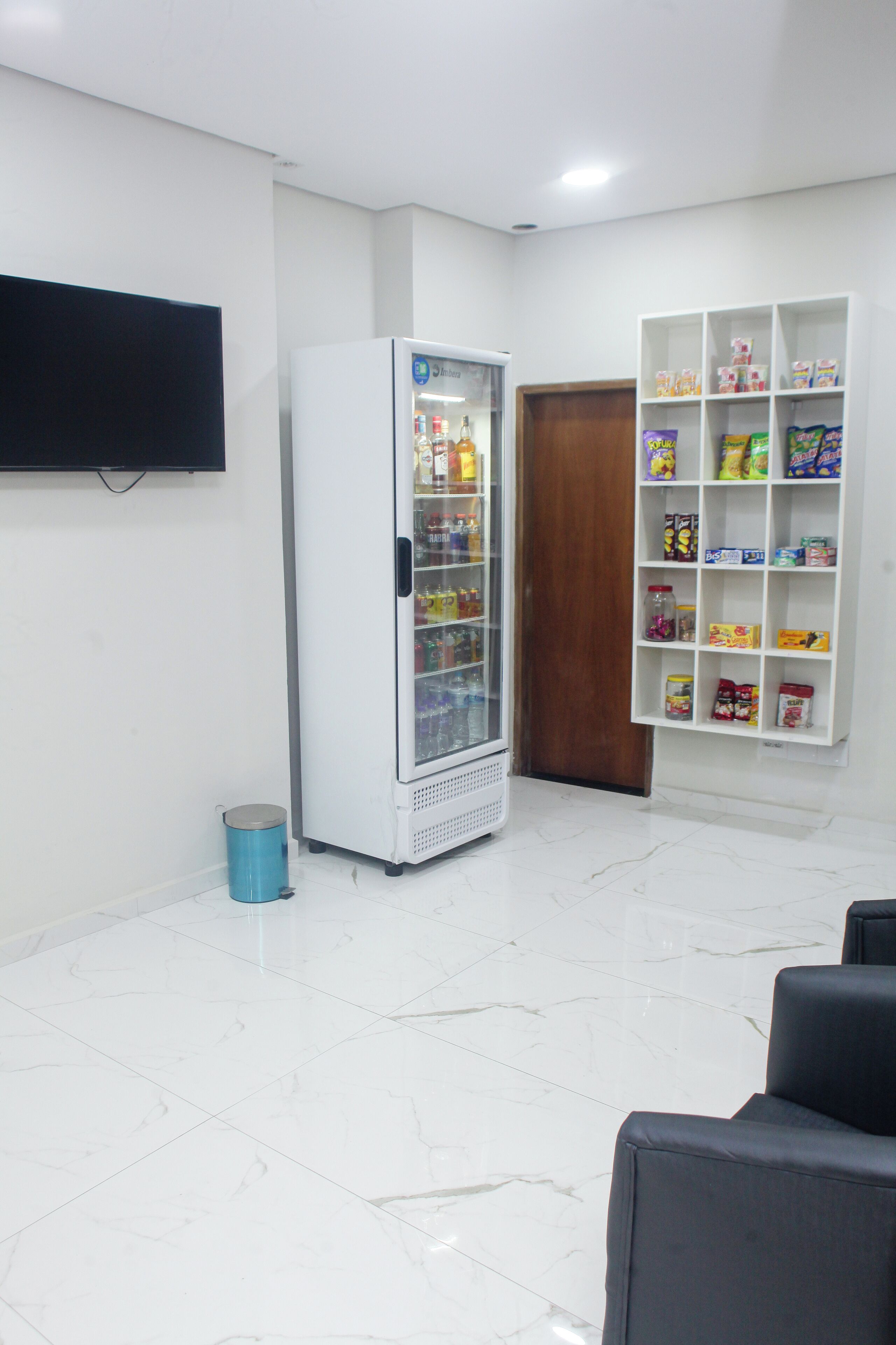 A room with a white refrigerator stocked with beverages, a blue trash bin, and a white shelving unit with snacks.