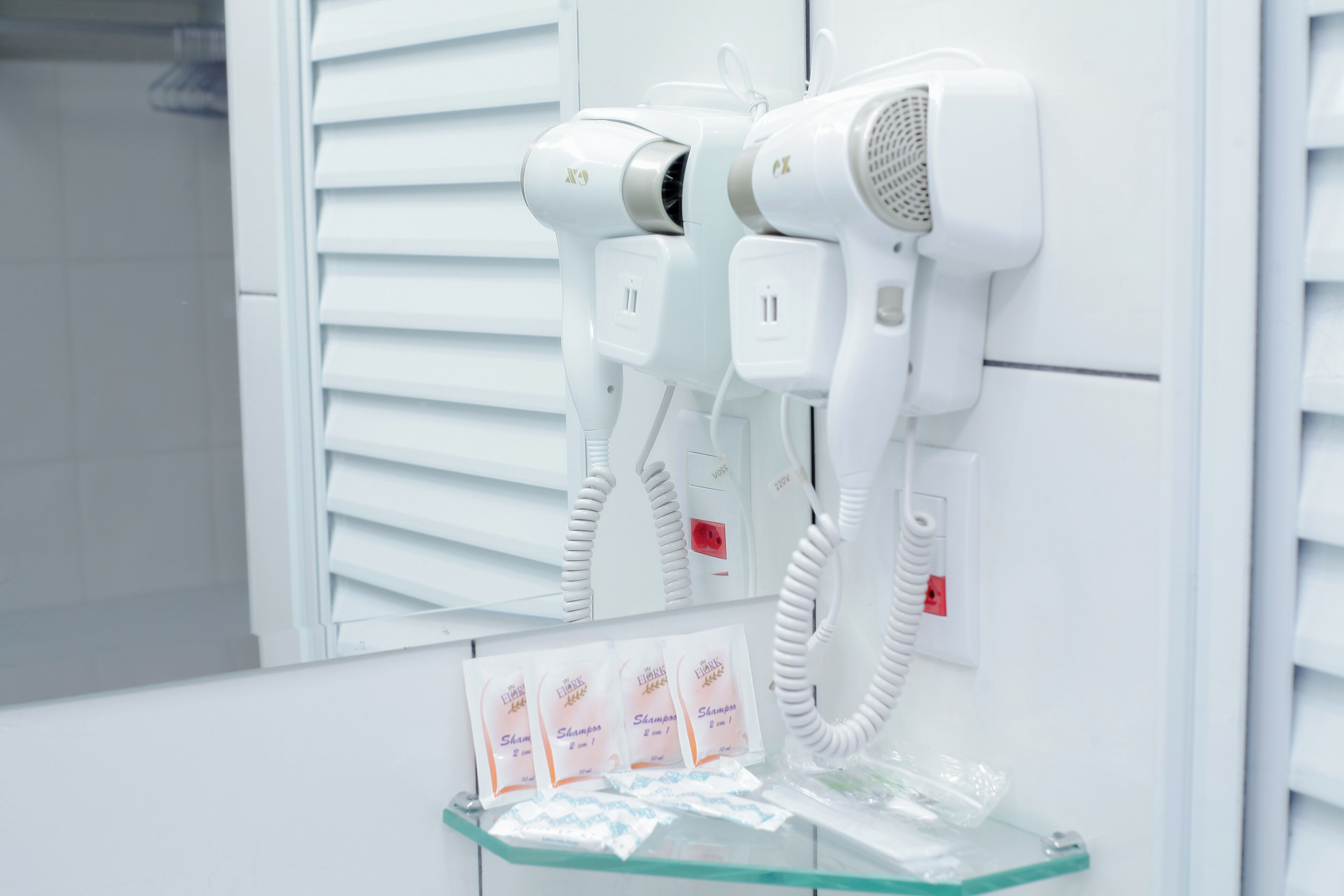 A bathroom with two white hair dryers mounted on the wall, a glass shelf with shampoo packets, and a white tiled floor.