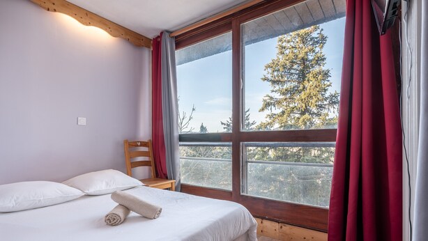 Un dormitorio con un ventanal grande, una cama con ropa de cama blanca, una silla de madera y una mesita.