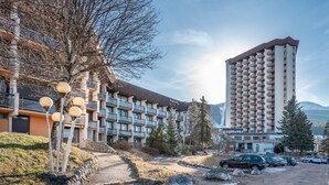 Un hotel de varios pisos con balcones, zona de estacionamiento y montañas al fondo.