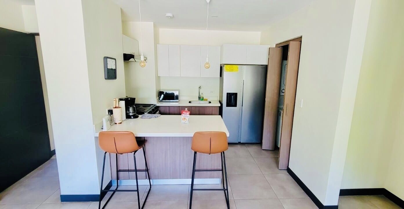 Una cocina compacta con una isla central, dos taburetes de bar, un refrigerador y un microondas.