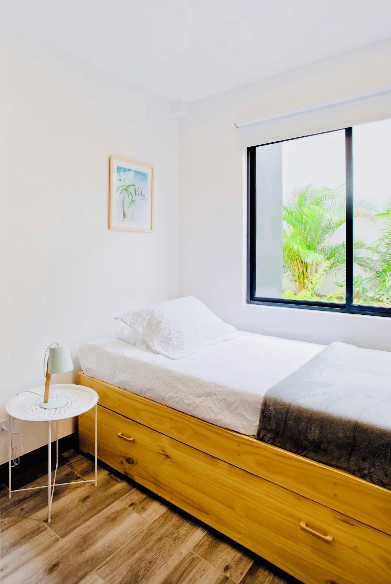 A bedroom with a bed, a small white table with a lamp, a wooden dresser, and a window with a view of greenery.