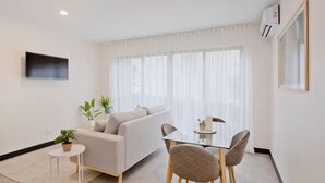 Una sala de estar moderna con un sofá, una mesa de comedor de vidrio y sillas. Hay una mesita auxiliar con una planta y un televisor montado en la pared.