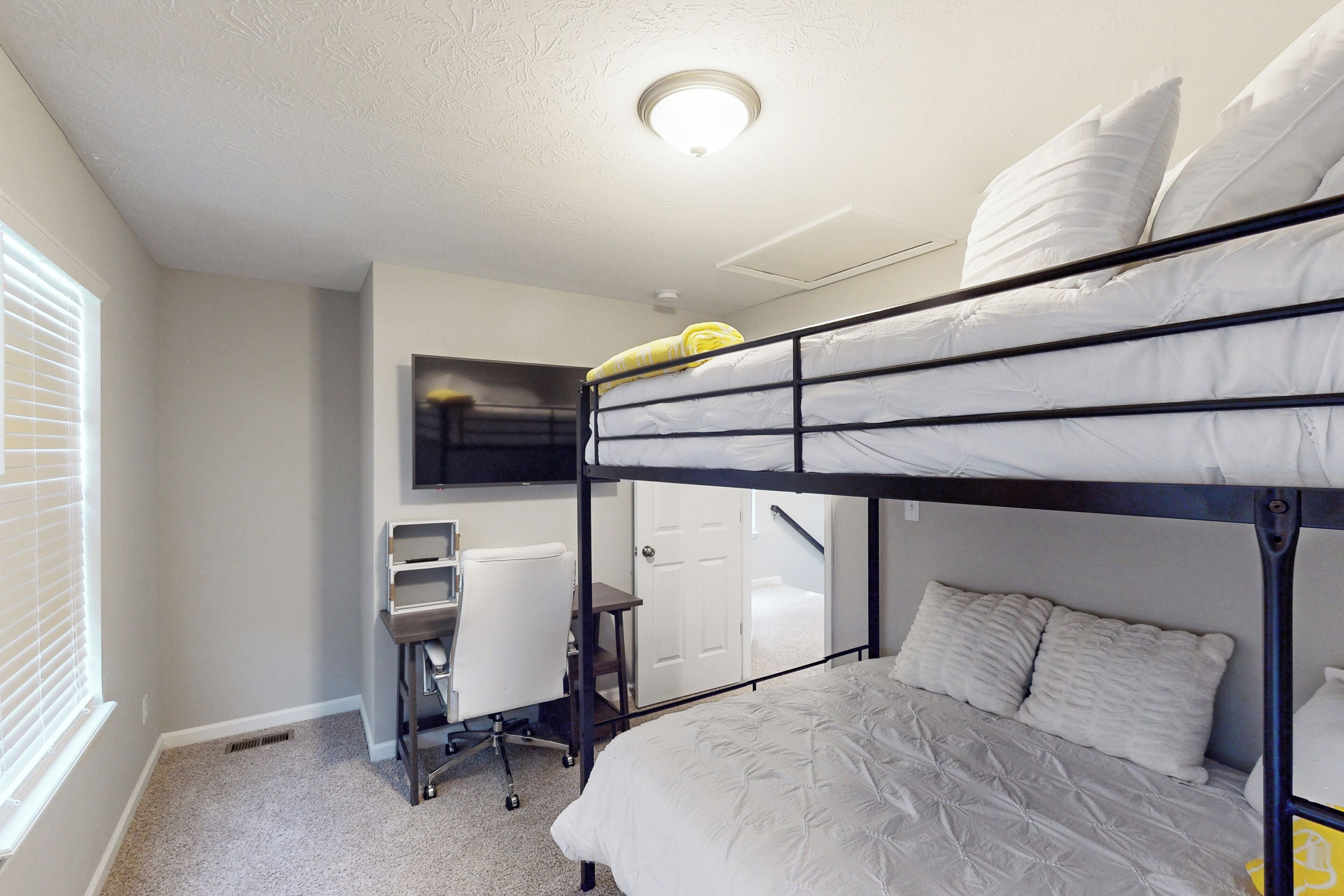 Une chambre avec un lit superposé, un bureau et une chaise, une fenêtre avec des stores et une porte donnant sur une autre pièce.