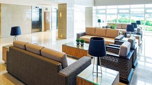 A spacious lobby with a modern design, featuring a seating area with sofas and a coffee table, a reception desk, and large windows offering a view of the outdoors.