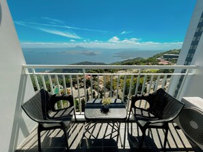 Un balcón con dos sillas y una mesa, con vista a un paisaje costero que incluye montañas y una extensión de agua.