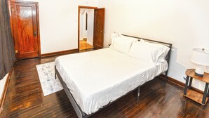 Un dormitorio con suelo de madera, una cama con ropa de cama blanca, una mesita de noche con una lámpara y una puerta de madera.