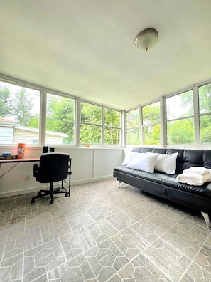 A room with a black leather sofa, a black office chair, a desk with a computer, and a window with a view of trees.