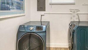 Uma lavanderia com duas máquinas de lavar de abertura frontal, uma porta, uma janela com persianas e um quadro na parede.
