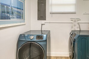 A laundry room with two front-loading washing machines, a door, a window with blinds, and a framed picture on the wall.