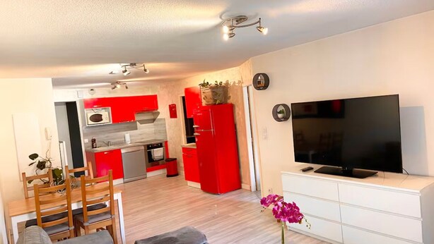 Un salon moderne avec un canapé gris, une table à manger en bois et des chaises. Un téléviseur à écran plat est fixé sur un meuble blanc. La cuisine est dotée d’appareils électroménagers rouges et d’un four à micro-ondes.