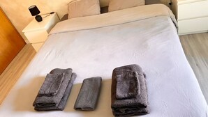 Un lit bien fait, recouvert d’une courtepointe blanche, avec trois serviettes pliées posées dessus. Il y a deux tables de chevet, chacune surmontée d’une lampe.