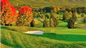 Un campo de golf con colinas onduladas, árboles con follaje otoñal y un cielo despejado.