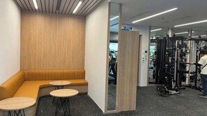 Un gimnasio moderno con una pared de madera, una zona de estar con sofá y mesas, y diverso equipo de ejercicio.