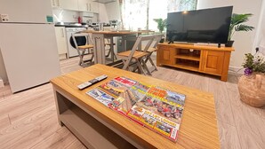 Een moderne woonkamer met een houten salontafel, een flatscreen televisie op een houten standaard, een eethoek met een tafel en stoelen, en een keuken op de achtergrond.
