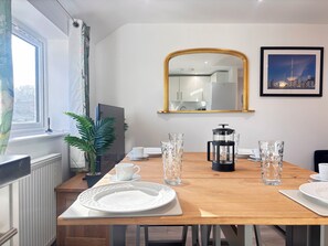Een eetgedeelte met een houten tafel gedekt voor vier personen, een koffiezetapparaat en een lijst met een stadsgezicht aan de muur.