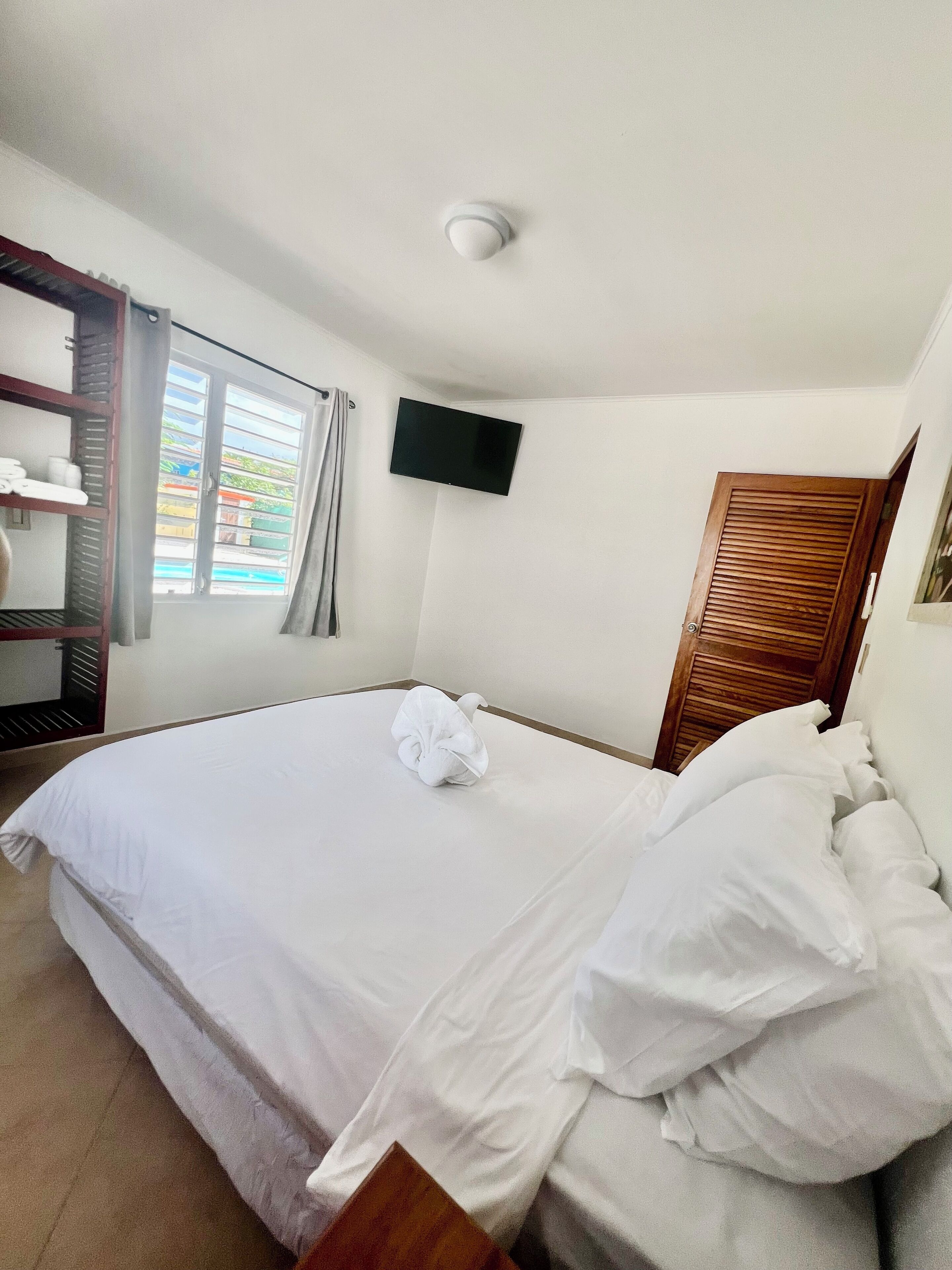 A hotel room with a bed, a flat-screen TV mounted on the wall, a wooden door, and a window with curtains.