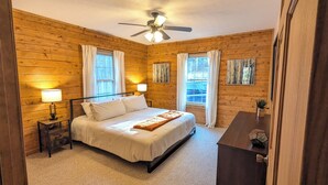 Ein Schlafzimmer mit Holzwänden, einem Bett, Nachttischlampen, einem Deckenventilator und zwei Fenstern mit Vorhängen.