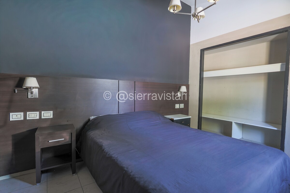 Une chambre à coucher comprenant un lit, des tables de chevet, une commode, une étagère fixée au mur et un luminaire au plafond.