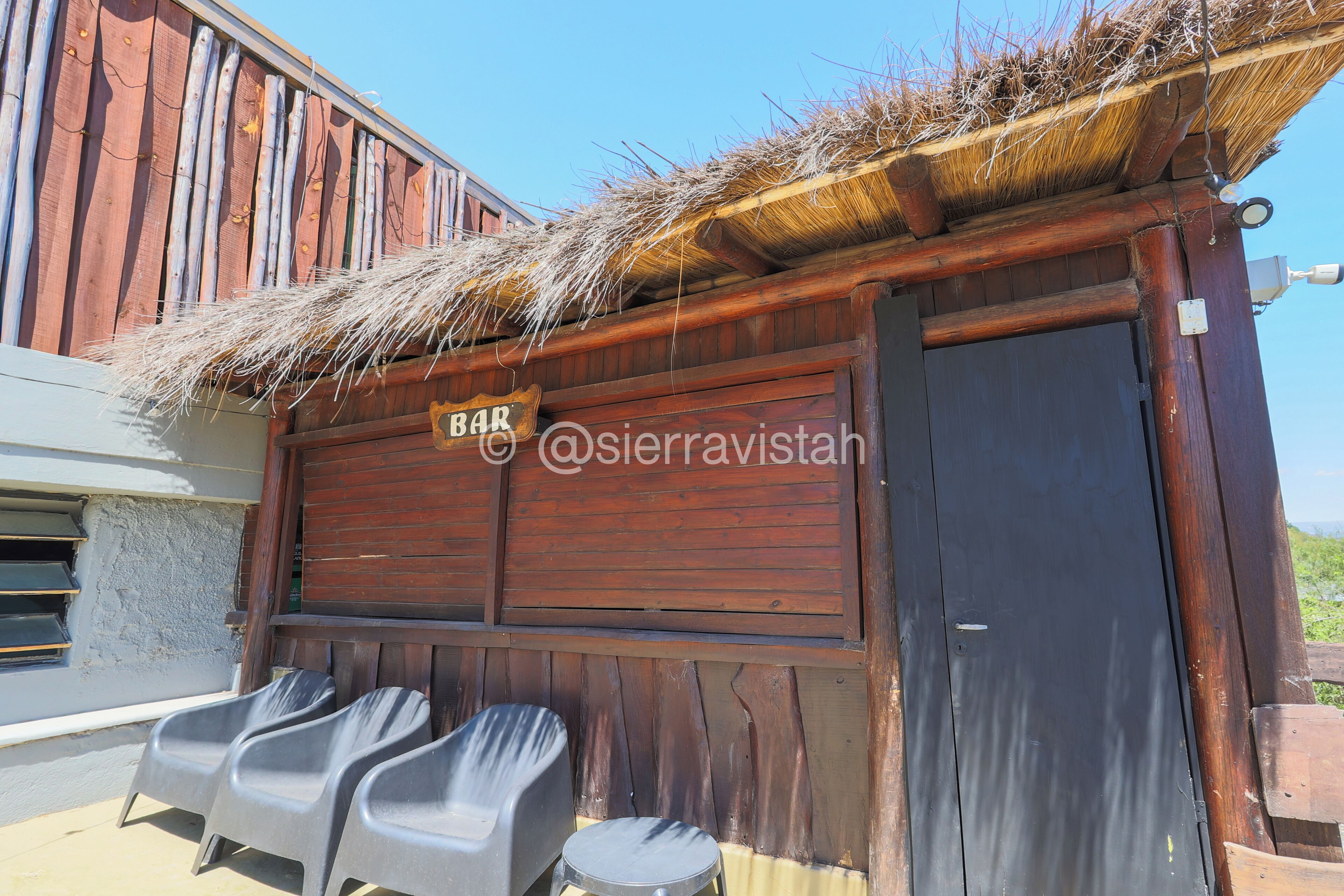 A rustic wooden building with a thatched roof, a sign reading "BAR", and plastic chairs outside.
