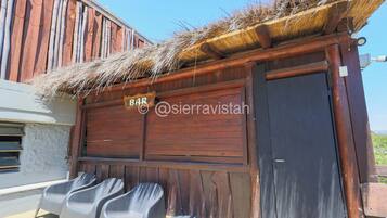 A rustic wooden building with a thatched roof, a sign reading "BAR", and plastic chairs outside.