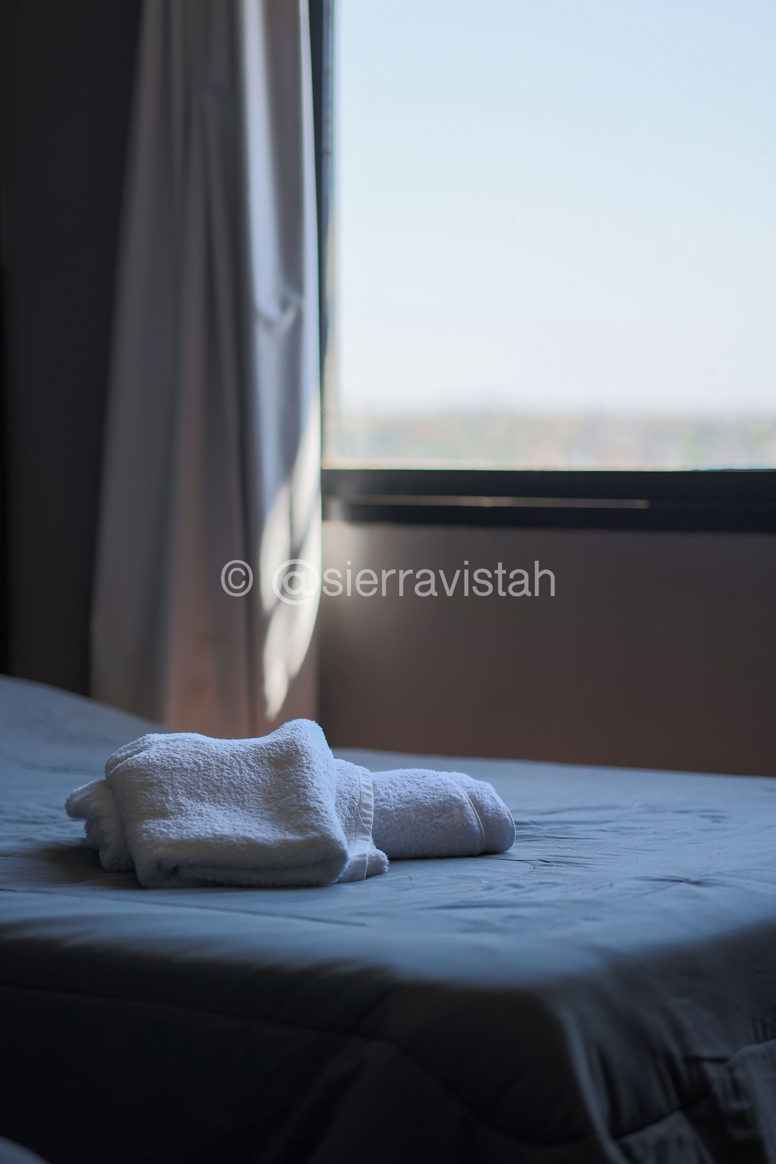 A neatly made bed with a folded white towel on it, a window with curtains, and a view of the sky.