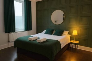 A hotel room with a bed, green curtains, a round mirror, a bedside lamp, and a radiator. - The Cathays Nook - Spacious 2 Bed Apartment Central Cardiff (Cardiff)