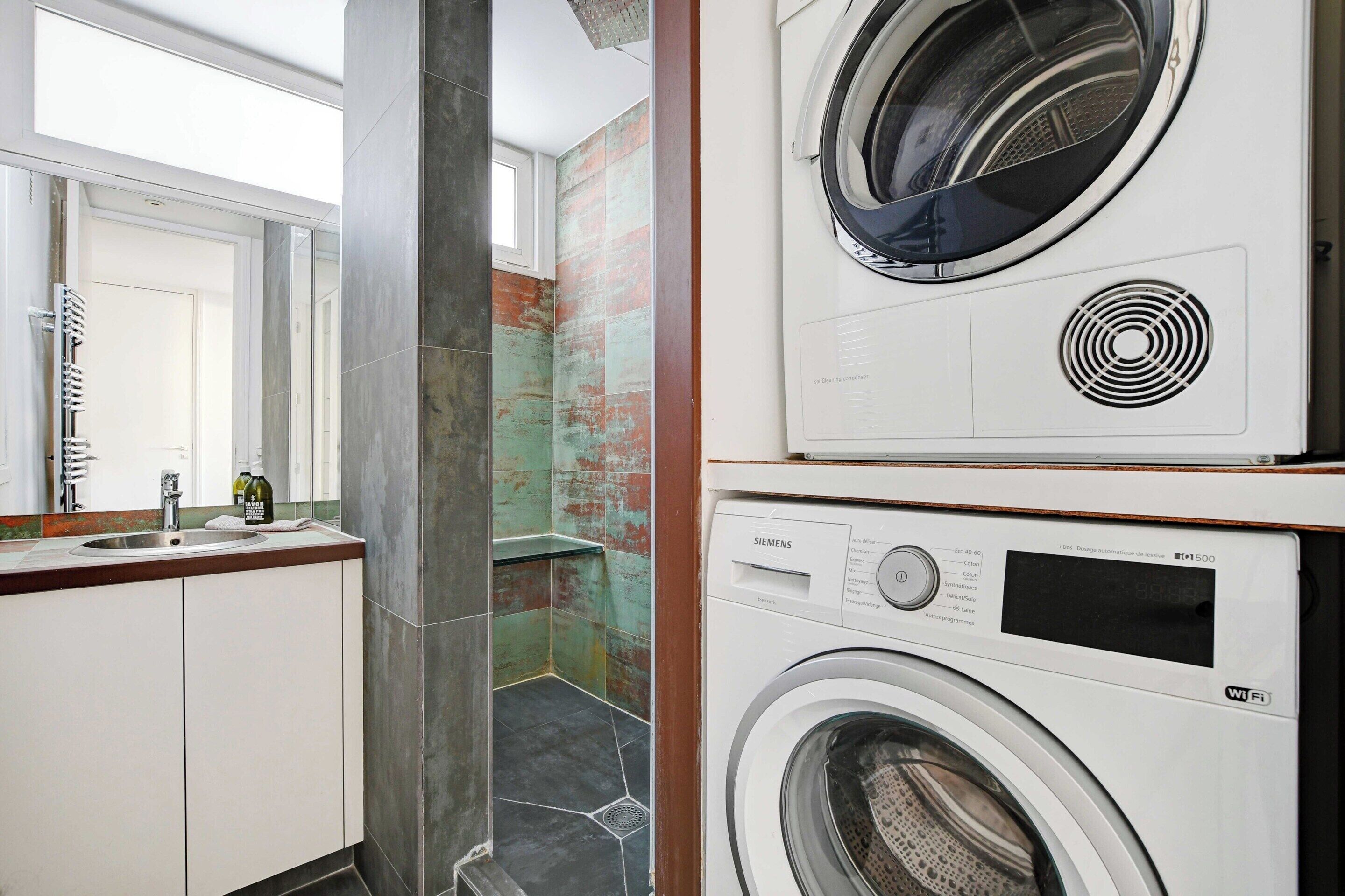 A modern laundry room with a Siemens washing machine and dryer, a sink with a mirror, and a view of a bathroom with a shower and a tiled wall.