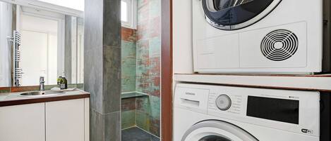 A modern laundry room with a Siemens washing machine and dryer, a sink with a mirror, and a view of a bathroom with a shower and a tiled wall.