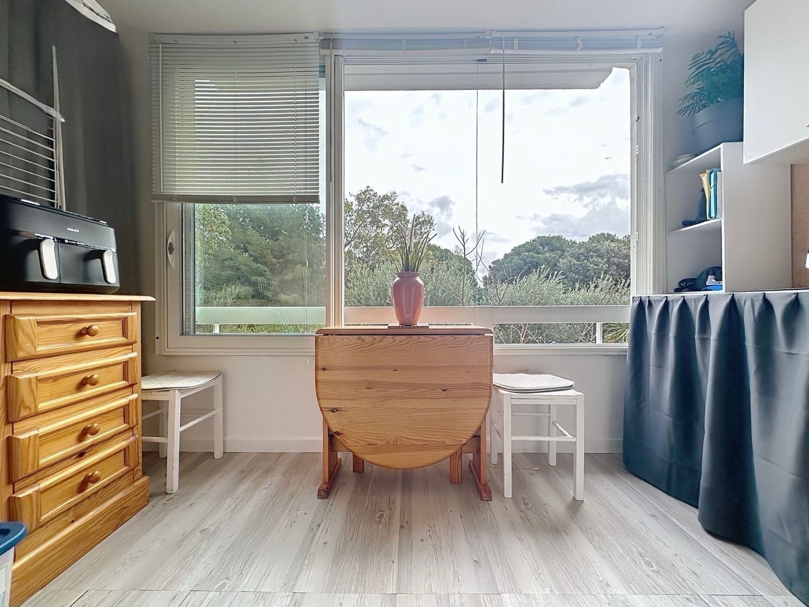 Una habitación con cómoda de madera, una mesita, una silla, un ventanal con persianas y vistas al exterior.