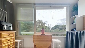 Une pièce comprenant une commode en bois, une petite table, une chaise, une grande fenêtre avec des stores et une vue sur la verdure à l’extérieur.