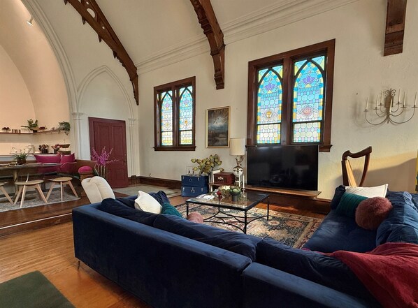 A spacious living room with a large blue sofa, a coffee table, and a television. There are two large stained glass windows and a wooden ceiling. - Unique Church Stay! Hot tub, River, Town (Savanna)