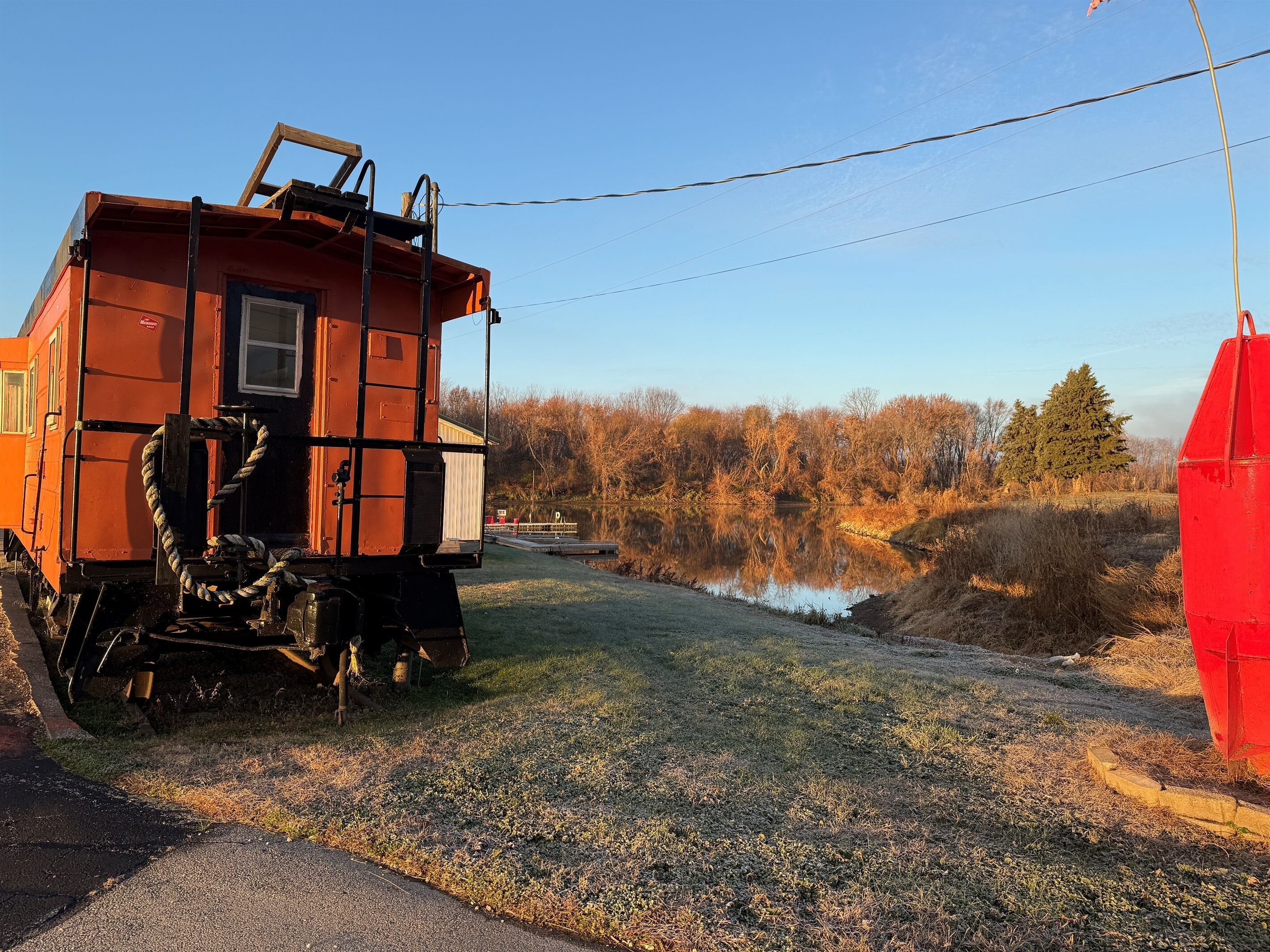 A train car with a ladder and a rope, parked beside a road with a grassy area and trees in the background.